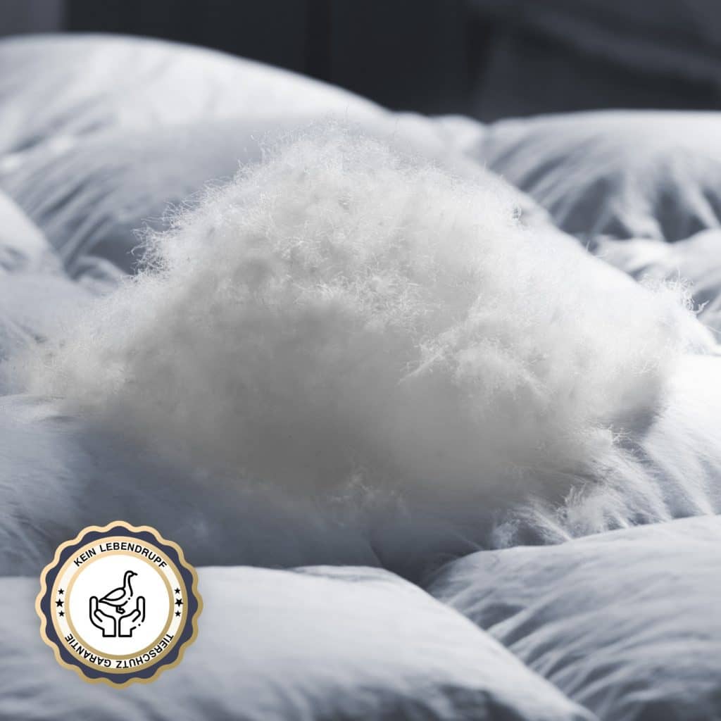 A fluffy white down cluster rests on a soft, quilted surface. A circular emblem with a hand icon and German text is at the bottom left corner, suggesting quality assurance typical of luxury down comforters.