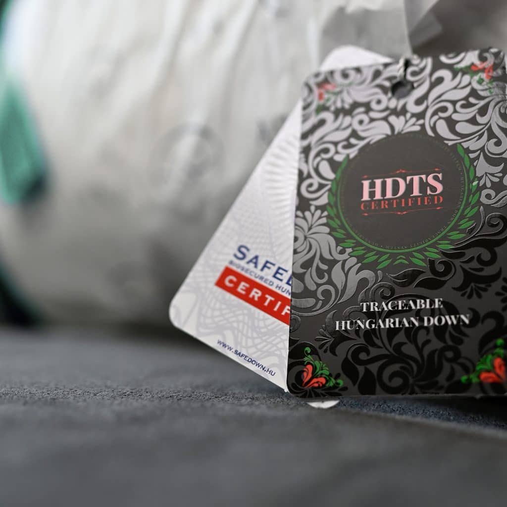 Close-up of product tags on Hungarian goose down duvets. The foreground tag reads HDTS Certified with intricate black and green designs, while another tag peeks out partially, showing Safe Certified. The background is softly blurred fabric.